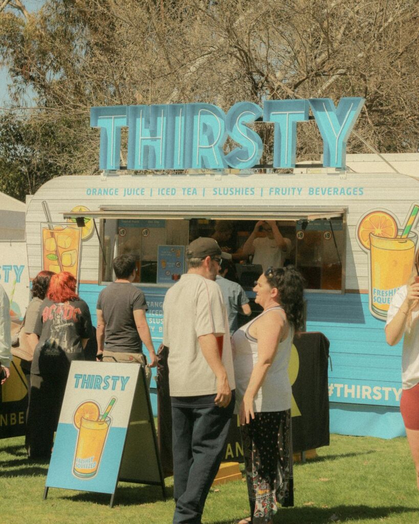 A white food truck with a light blue sign atop it that says thirsty. People are lined up to buy drinks.